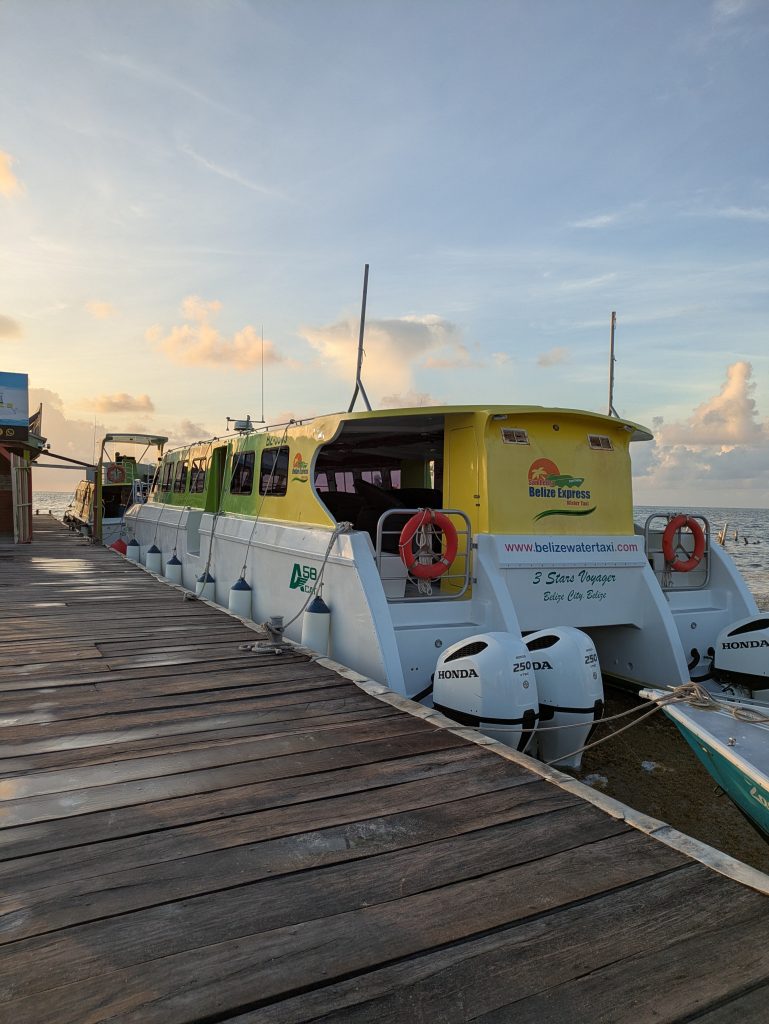Photo of a dock with two boats tied up on the right hand side. They are both white on the bottom with yellow fading into green on the top. The front one has the words "San Pedro Belize Express Water Taxi" on it, with a link to the website underneath. The ship is called 3 Stars Voyager. The ferry in front of it is the Caye Caulker to Mexico one and it is smaller but same colours. On the left you can make out a building with part of the roof visible.