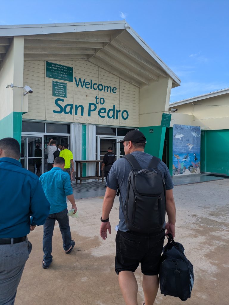 Photo showing multiple passengers walking towards a large building that says Welcome to San Pedro. There is another sign that is blurry but says it is the San Pedro Port of Entry.