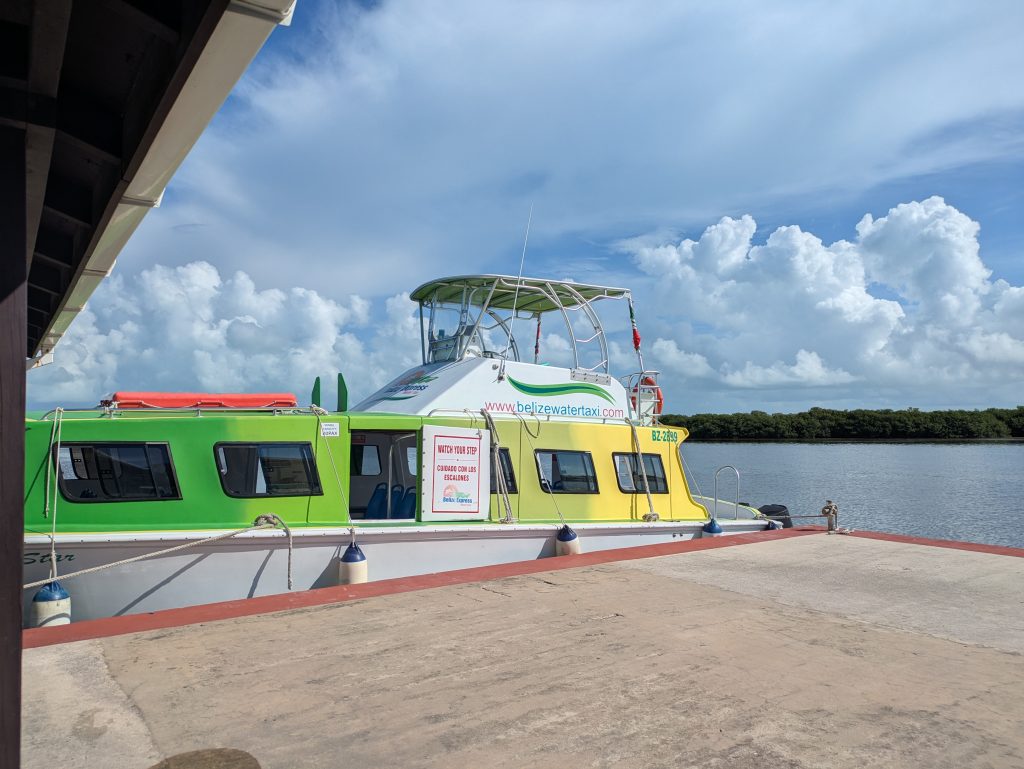 Photo of the Caye Caulker to Mexico ferry. It is yellow on the right fading to green on the left. It is docked with a large empty concrete space. In the back on the other side of the water are lots of trees.