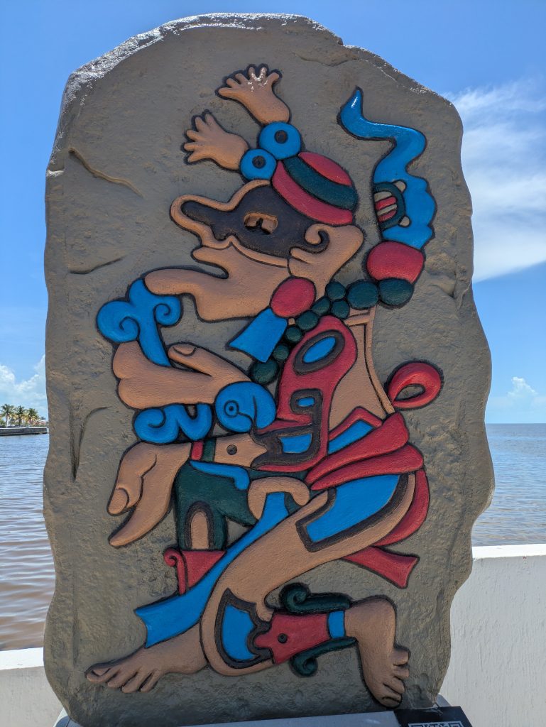 Photo of a stone featuring a colourful Mayan carving of a deity. The carving is painted using lots of bright red and blue colours. The figure is down on one knee.