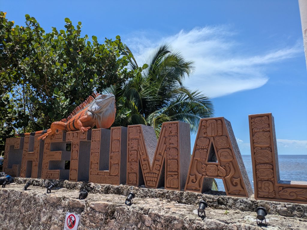 Photo of a sign that says Chetumal. There is a statue of a lizard on the top of the sign. Each letter is decorated with lots of Mayan designs. Behind the sign are some trees and the sea.