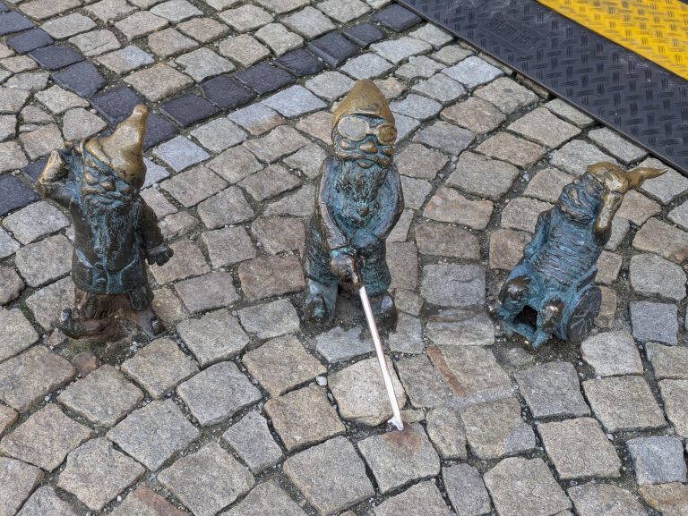 Photo of three dwarf statues. One is holding a hand to their ear, one is holding a white stick in front of them and one is in a wheelchair.