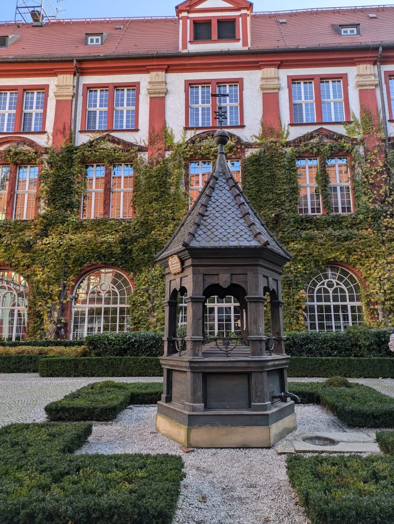 Photo of a water fountain in front of a building containing large windows and covered in ivy. The top floor is clear of Ivy and you can make out red columns between each window.